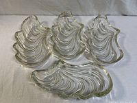 Four clear glass dishes shaped like seashells with swirling wave patterns on a white cloth background.