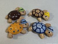 Five hand-painted ceramic turtle figurines arranged on a flat surface, showing various shell colors and hats.