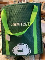 Dark green insulated reusable grocery bag branded T&T with bright green handles and white logo of a smiling dumpling.