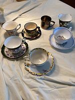 Seven cups and saucers, mostly bone china, with floral patterns and one metal cup without saucer, laid out on cloth