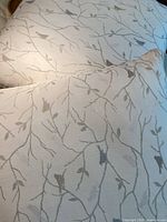 Two neutral decorative cushions with embroidered branches, leaves, and birds, displayed on a cream background fabric.