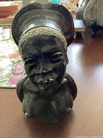 Black stone statue showing detailed female head sculpture with headdress, carved from heavy black stone.