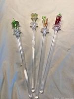 Four clear plastic drink stirrers with decorative tops shaped like a green bottle, diamond, carrot, and berry cluster.