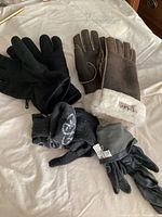 Four pairs of gloves displayed on a bed, including black leather, brown suede with shearling, black fabric, and knitted gloves.