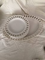 Full set of 6 stacked white ceramic plates and one matching oval serving platter arranged on white fabric background.