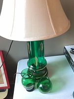 Green glass lamp with gold leaf detailing and beige fabric shade with two smaller matching green glass vases with gold trim placed in front on a white surface.
