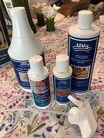 Four bottles of UV3 leather and wood care products with white caps and blue labels, arranged on a floral tablecloth along with a white spray nozzle.