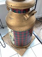 Close-up photo of a large vintage metal milk container with a plaid fabric decoration wrapped around the midsection, mounted on a wooden base with a metal handle.