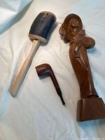 Three items displayed on a cloth: carved wooden female figure, old smoking pipe, and wooden noise maker.