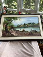 Framed oil painting showing a detailed landscape with a lake, trees, houses, and people fishing.