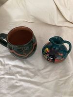 Photo of a dark green ceramic cup with reddish-brown interior and a teal jug with tropical painted scene.