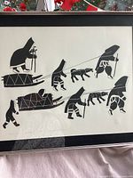 Framed black and white indigenous print showing Inuit figures with sleds and dogs in silhouette style.