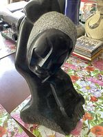 Full view of the black and gray stone sculpture showing the stylized female face resting on her hand.