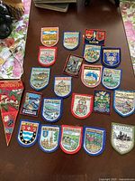 Photo showing 19 assorted embroidered travel badges from England and Wales laid out on a table, each with various scenic and heraldic designs and text of place names.