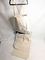 Photo showing multiple blank canvas tote bags stacked with one hanging on hanger, displaying the undyed off-white fabric and sturdy strap.