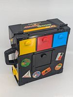 Black Fisher Price Magic Show In A Box toy with three colorful drawers (yellow, red, blue) and various travel-themed stickers on front and sides.