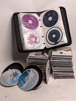 Photo of the black CD binder open with multiple CDs in plastic sleeves and two stacks of CDs beside it, showing a variety of discs and album art.
