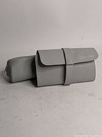 Full view of the closed fold-over makeup clutch and cylindrical zippered pouch, showing grey pleather exterior and brand embossing.
