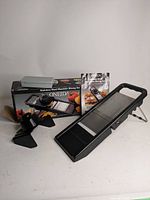 Photo showing the mandolin slicer with its foldable stand, original box, and instruction manual.