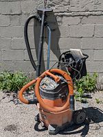 Front view of Rigid wet/dry shop vacuum with attached hoses and cables, showing orange tank body and black lid.