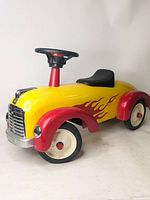 Side view of yellow and red metal ride-on toy car with flame decals and black seat, steering wheel, and cream wheels.