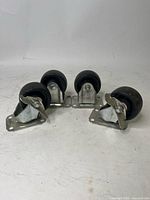 Four caster wheels, two swivel and two fixed, with metal plate mounts and black plastic wheels, shown arranged on white background.