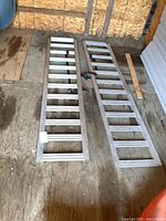 Two aluminum loading ramps laid side by side on a wooden floor, showing the ladder rungs and flat base.