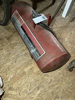 Full view of the red Sanborn portable air tank resting on a wooden floor, showing the shape and handle.