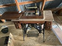 Antique Singer sewing machine on wooden table with cast iron treadle base, viewed from front and side angles showing details of machine, cabinet drawers, and treadle mechanism.