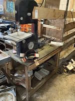 Full side view of the black Shopmaster bench top bandsaw mounted on a wooden base with spare bandsaw blades coiled on the base shelf, showing electric motor and belt drive mechanism.