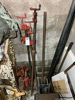 Photo showing three red bar clamps and a rusty trolley jack leaning against a concrete wall, with metal tubes and brushes nearby.