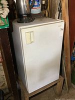White Sanyo bar fridge standing on a wooden stool, with some items on top, in a storage or workshop area.