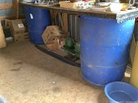 Pair of large blue plastic barrels in indoor setting with surrounding items and floor visible.