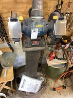 Front view of Mastercraft 8 inch bench grinder mounted on stand, showing grinding wheels, protective shields, and manual on base.