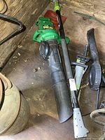 Full view of the green Weed Eater Barracuda electric leaf blower and Earthwise electric pole saw with black extension pole on wooden floor, showing condition and accessory tubes.