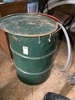 Green steel burn barrel with removable lid, visible dents, and attached transparent hose coiled around it.