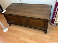Wooden cedar chest with carved decorative panel on the front and flat top surface, showing overall view of item.