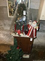 Two 32 inch artificial Christmas trees, one decorated and covered in plastic, the other undecorated; two plush Santa figures; and a wooden surface holding candle holders.