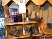 Small vintage glass bottles, cardboard tobacco pack, small copper and brass bugle, metal bell, figurine and other small collectibles on wooden shelf.