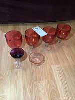 Four large cranberry glass goblets, one amethyst glass goblet, and one small pink glass nut bowl displayed on wooden floor.