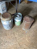 Photo showing all three cans: a large vintage steel cream can with rusted lid, a small farmhouse-tin can in the middle, and a tall rusted metal can on the right