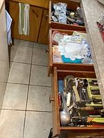 Three kitchen drawers displaying contents: a drawer with cloth towels and doilies; a drawer containing assorted cutlery and kitchen utensils; another drawer with additional kitchen tools, small tools and cutlery.