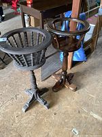 Photo showing two different solid wood fern stands side-by-side on a plywood floor in a storage area.