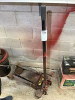 Overall side and handle view of the hydraulic floor jack leaning against a concrete wall.