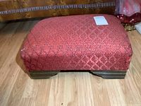 Front-side view of the antique wooden footstool showing red patterned upholstered top with some fabric wear.