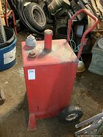 Full view of a red metal fuel tank with handle and wheels, showing wear and surface rust.