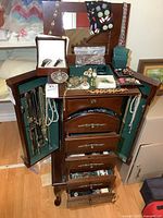 Open wooden jewelry cabinet showing necklaces hanging inside doors, drawers open with bracelets, earrings, small boxes and crystal dish visible