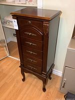 Front and side view of the closed jewelry cabinet showing wood finish, 5 drawers with ornate metal handles, decorative carved details on the corners, and cabriole legs.