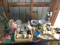 Wide view of wicker tray with costume jewelry, decorative dishes, candle holder, vase, and other assorted collectibles on a table with a corrugated metal background.