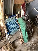 View of five lawn chairs including three macrame woven metal frame chairs stacked, a camp chair in a green bag, and a wooden folding chair leaning against the wall.